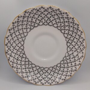 May include: A white saucer with a gold rim and a black and white lattice design. The saucer is made of fine china and has a delicate scalloped edge.