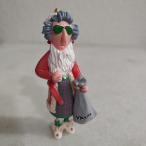 May include: A figurine of a woman wearing green sunglasses, a red coat, and a green and red plaid skirt. She has a white beard and is holding a gray bag with the name "Maxine" written on it. The figurine is wearing white bunny slippers.