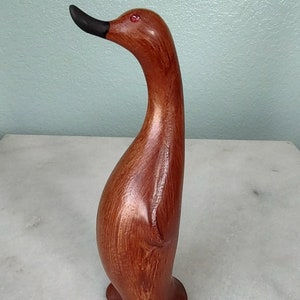 May include: A wooden carved penguin figurine with a long neck and red eyes. The penguin is painted a reddish-brown color.