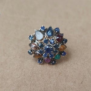 Pode incluir: Um anel de ouro com um grupo de pedras preciosas coloridas, incluindo safiras azuis, rubis vermelhos, esmeraldas verdes e olhos de tigre marrons.