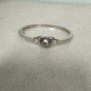 Puede incluir: Un brazalete de plata con una sola perla gris y detalles de cristal transparente.