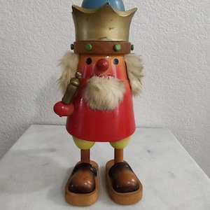 Puede incluir: Una figura de cascanueces de madera con un cuerpo rojo y amarillo, una barba blanca y una corona azul y dorada. El cascanueces sostiene una pipa dorada en su mano derecha y lleva zapatos de madera marrones.