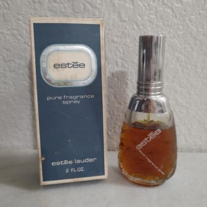 Op de afbeelding: Een blauwe en witte doos met de tekst "Estée Lauder" en "pure fragrance spray". De doos bevat een heldere glazen fles met parfum en een zilveren dop. De fles heeft de tekst "Estée" en "pure fragrance spray". De fles is 2 fl oz.