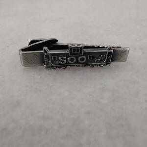 May include: A silver tie clip featuring a detailed model of a train engine with the text "SOO 67" on the side.