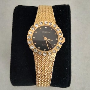 May include: A gold-tone wristwatch with a black face and a crystal-encrusted bezel. The watch face has the word "CYRANO" printed on it and says "QUARTZ" below the 12 o'clock position.