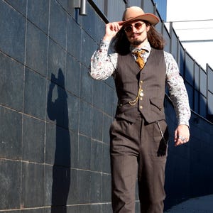 Puede incluir: Un chaleco y pantalones de rayas marrones combinados con una camisa de manga larga con estampado floral, un sombrero fedora color canela, gafas de sol redondas, una cadena de reloj de bolsillo dorada y botas de cuero marrón.