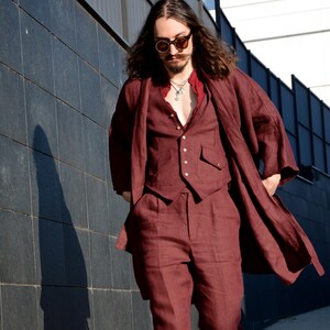Può includere: Un completo in lino bordeaux composto da un cappotto lungo, un gilet e pantaloni. L'abito è abbinato a una camicia rossa e scarpe in pelle marrone. L'outfit è indossato all'aperto, con un muro scuro piastrellato sullo sfondo.