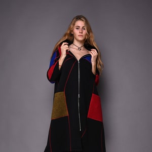 Op de afbeelding: Een lange zwarte jas met een ritssluiting over de hele lengte, met kleurvlakken in rood, blauw en olijfgroen. De jas heeft rode stiksels en wordt gedragen met zwarte laarzen.