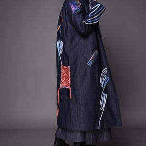 Puede incluir: Un abrigo largo de mezclilla azul oscuro con detalles bordados coloridos y parches. El abrigo presenta diseños abstractos blancos y azules, y un parche rojo texturizado. Las mangas tienen detalles en blanco y azul. El abrigo se usa con botas negras.