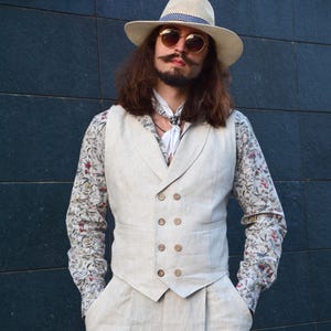 Puede incluir: Un chaleco cruzado de color beige y pantalones a juego combinados con una camisa de manga larga con un estampado floral en rojo, verde y gris. El conjunto incluye un sombrero de paja con una banda azul estampada, gafas de sol redondas y un pañuelo blanco.