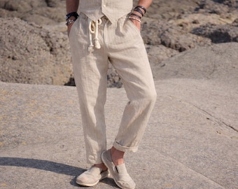 May include: Beige linen trousers featuring a drawstring waist with thick white rope ties, side pockets, and a slightly tapered leg with a cuffed hem. The pants are paired with a matching beige button-up vest and light-colored slip-on shoes.