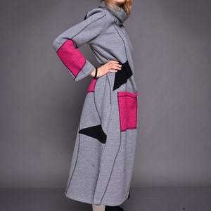 Op de afbeelding: Een grijze jurk met lange mouwen en een hoge kraag, met roze en zwarte geometrische patches. De jurk heeft een knielange rok en wordt gedragen met zwarte laarzen.