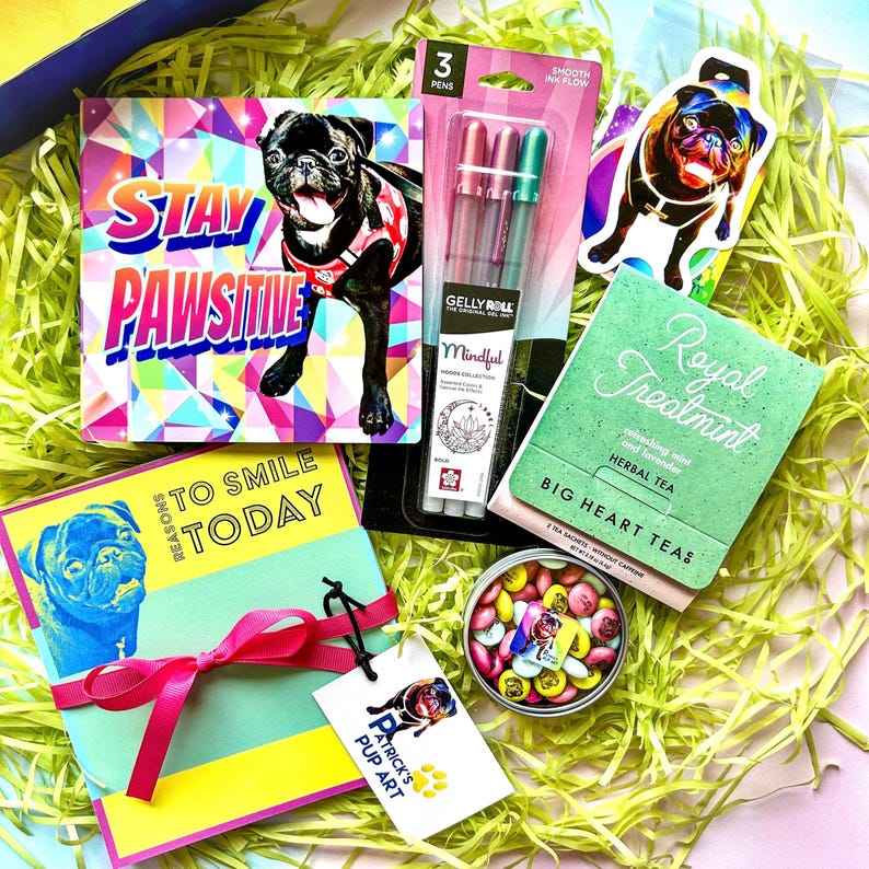Op de afbeelding: Een cadeaudoos gevuld met artikelen voor een hondenliefhebber. De doos bevat een kaart met de tekst "Stay Pawsitive", een kaart met de tekst "Reasons to Smile Today", een pak met drie gelpennen, een sticker van een mops, een blikje snoep en een doos kruidenthee.