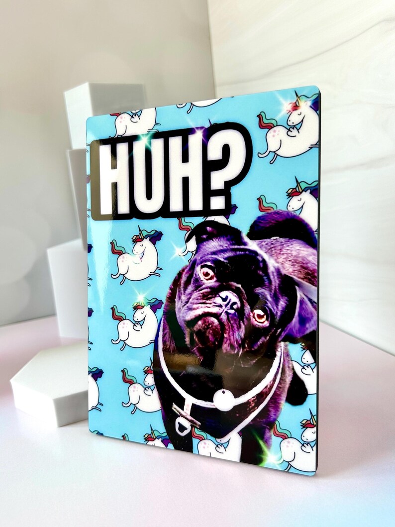 K&ouml;nnte beinhalten: Ein schwarzer Mops mit einem wei&szlig;en Halsband schaut in die Kamera. Der Hund befindet sich auf einem blauen Hintergrund mit einem Muster aus wei&szlig;en Einh&ouml;rnern. Der Text "HUH?" ist in wei&szlig;en Buchstaben auf schwarzem Hintergrund.
