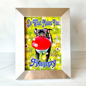 Puede incluir: Una impresi&oacute;n enmarcada con una imagen colorida de un perro pug negro atrapando un frisbee rojo en un campo de hierba verde con flores rosas. El texto "Do What Makes You Happy" est&aacute; escrito en letras azules en la parte superior de la imagen.