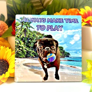 May include: A black pug dog with a playful expression is holding a purple and yellow ball in its mouth. The dog is standing on a beach with palm trees and blue sky in the background. The text "ALWAYS MAKE TIME TO PLAY" is written in blue and white letters on the top of the image.