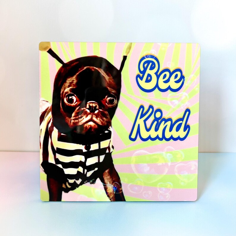 May include: Square print featuring a black pug in a bee costume with antennae and a striped shirt. The background has pink and green stripes with the words "Bee Kind" in a playful font.