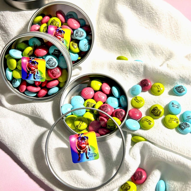 Op de afbeelding: Drie zilveren blikken gevuld met kleurrijke M&Ms snoepjes. De M&Ms zijn versierd met een mopshond ontwerp en de woorden "Pug's Pup Art".