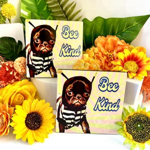 May include: Two square decorative tiles with a pug wearing a striped shirt and a bee-themed hat. The tiles have the text "Bee Kind" in blue script on a yellow and pink background. The image is surrounded by yellow and orange flowers.