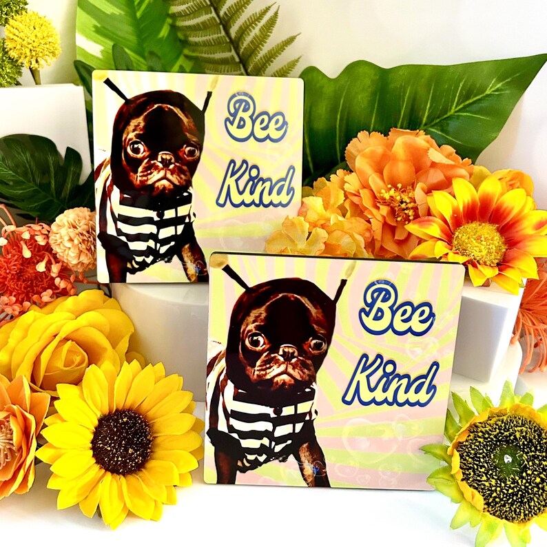 May include: Two square decorative tiles with a pug wearing a striped shirt and a bee-themed hat. The tiles have the text "Bee Kind" in blue script on a yellow and pink background. The image is surrounded by yellow and orange flowers.
