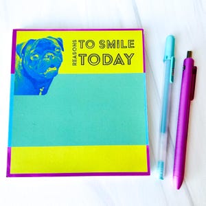 Op de afbeelding: Een geel en turquoise notitieboekje met een paarse rand. Het notitieboekje heeft een afbeelding van een mopshond en de tekst "Reasons to Smile Today". Twee pennen, een turquoise en een paarse, liggen naast het notitieboekje.