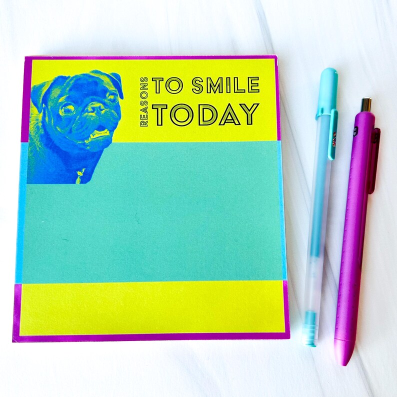 Op de afbeelding: Een geel en turquoise notitieboekje met een paarse rand. Het notitieboekje heeft een afbeelding van een mopshond en de tekst "Reasons to Smile Today". Twee pennen, een turquoise en een paarse, liggen naast het notitieboekje.