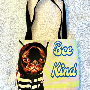 May include: A tote bag with black handles. The bag displays a pug wearing a striped shirt and a black hood. The words "Bee Kind" are written in blue and yellow on a pink and yellow background.