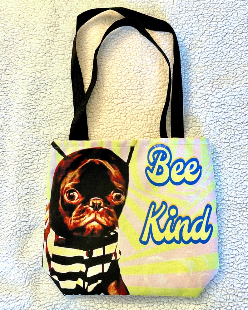 May include: A tote bag with black handles. The bag displays a pug wearing a striped shirt and a black hood. The words "Bee Kind" are written in blue and yellow on a pink and yellow background.