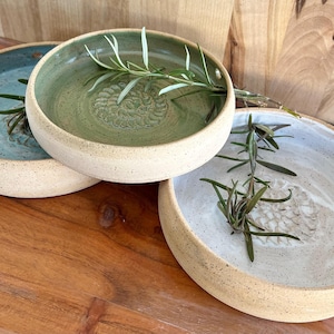 May include: Three round ceramic dishes with speckled beige outer walls and smooth, glazed interiors in shades of sage green, light blue, and white. Each dish features a textured, circular grater pattern at the center and a sprig of rosemary.