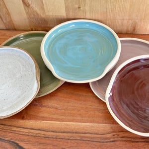 May include: A collection of five round ceramic spoon rests featuring a variety of glazed finishes including speckled white, olive green, bright turquoise, muted mauve, and deep burgundy, each with a raised edge and a small notch for a handle.