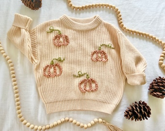 May include: A cream-colored knitted sweater with embroidered pumpkins in shades of brown and green.