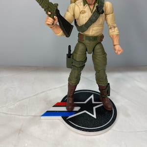 May include: A plastic action figure of a soldier with a green and black rifle, wearing a tan shirt, green pants, and brown boots. The figure is standing on a black and white star-shaped base with a red, white, and blue stripe.