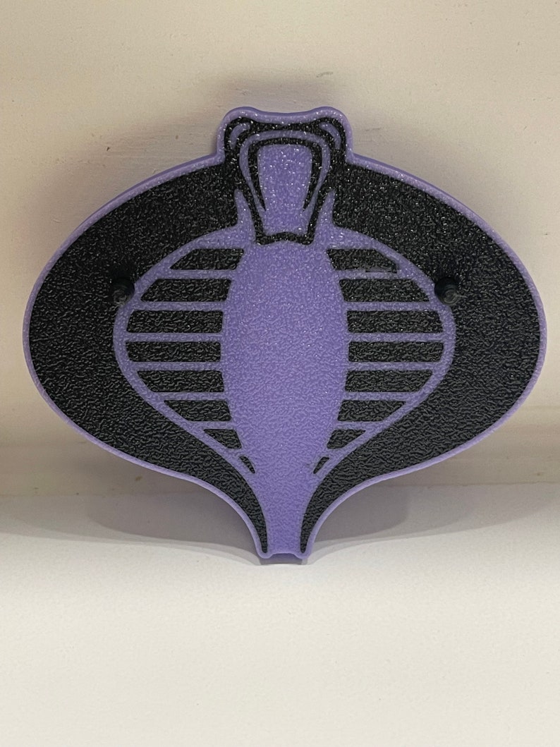 Peut inclure: Un embl&egrave;me Cobra en plastique violet avec un logo Cobra noir. Le logo Cobra est un serpent stylis&eacute; avec un capuchon sur&eacute;lev&eacute; et une s&eacute;rie de lignes horizontales sur son corps.