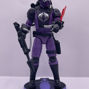 Peut inclure: Une figurine d'action violette d'un Cobra Trooper de la franchise G.I. Joe. La figurine tient un fusil et arbore un logo Cobra sur sa poitrine. La figurine est pos&eacute;e sur une base violette avec un logo Cobra.
