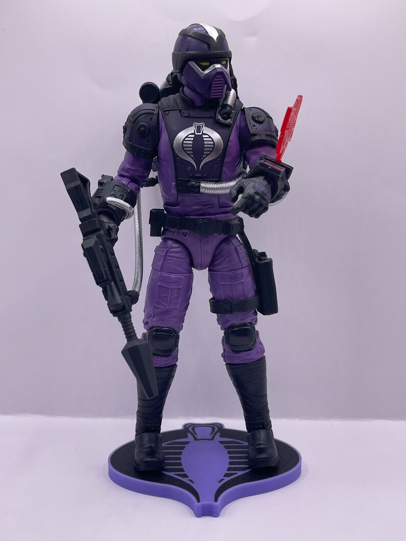 Peut inclure: Une figurine d'action violette d'un Cobra Trooper de la franchise G.I. Joe. La figurine tient un fusil et arbore un logo Cobra sur sa poitrine. La figurine est pos&eacute;e sur une base violette avec un logo Cobra.