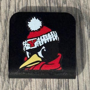 Peut inclure: Un charm carré noir présentant le dessin d'un pingouin portant un bonnet en tricot rouge et blanc avec un pompon et une écharpe rouge. Le bonnet affiche une lettre Y rouge sur le devant. Le pingouin a un bec jaune sur fond noir uni.