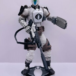 Peut inclure: Une figurine d'action blanche tenant un fusil noir et argent, une cha&icirc;ne noire et argent et une tron&ccedil;onneuse noire et argent. La figurine porte un casque blanc avec une visi&egrave;re bleue et un costume blanc et noir. La figurine est debout sur une base blanche avec un logo Cobra noir.