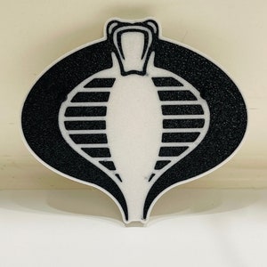 Peut inclure: Un embl&egrave;me de Cobra noir et blanc avec une t&ecirc;te de serpent et un corps ray&eacute;. L'embl&egrave;me est sur un fond blanc.