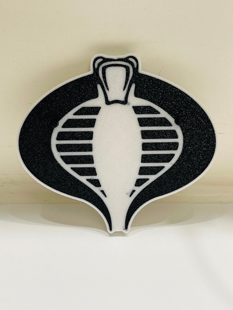 Peut inclure: Un embl&egrave;me de Cobra noir et blanc avec une t&ecirc;te de serpent et un corps ray&eacute;. L'embl&egrave;me est sur un fond blanc.