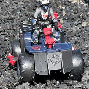 Peut inclure: Un quad jouet bleu et noir avec un logo Cobra à l'arrière. Une figurine d'action jouet portant un casque blanc et une armure grise et bleue conduit le quad.