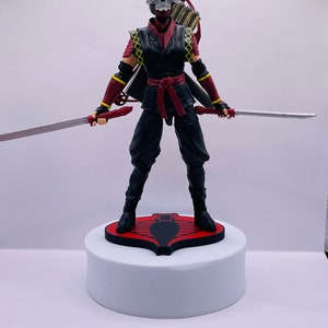 Peut inclure: Une figurine d'action noire et rouge d'un guerrier ninja avec un masque de cr&acirc;ne, tenant deux &eacute;p&eacute;es et un arc et des fl&egrave;ches. La figurine est debout sur une base rouge avec un logo de cobra noir.