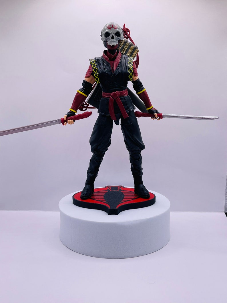 Peut inclure: Une figurine d'action noire et rouge d'un guerrier ninja avec un masque de cr&acirc;ne, tenant deux &eacute;p&eacute;es et un arc et des fl&egrave;ches. La figurine est debout sur une base rouge avec un logo de cobra noir.