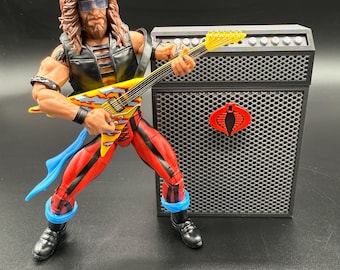 May include: Action figure of a rock star playing a yellow and red guitar, with a black amp featuring a red Cobra logo. The figure wears red pants with black stripes, a black vest, and blue accents. The figure has long brown hair and sunglasses.