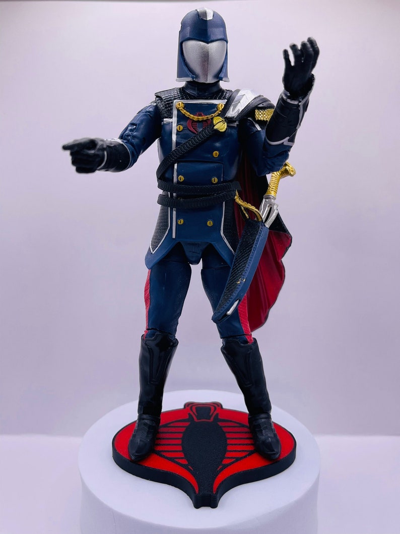 Peut inclure: Une figurine d'action de Cobra Commander, habill&eacute;e d'un uniforme bleu et argent avec une cape rouge et tenant une &eacute;p&eacute;e. La figurine est debout sur une base avec le symbole Cobra rouge et noir.