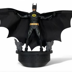 Può includere: Una action figure di Batman nera con un simbolo di pipistrello giallo sul petto, in piedi su una base nera con statue di gargoyle. La figura ha un mantello nero con ali di pipistrello.