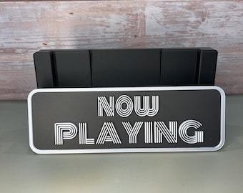 May include: A black sign with white text that reads "NOW PLAYING" in a retro font style. The sign is made of a thick, durable material and has a slightly raised edge.