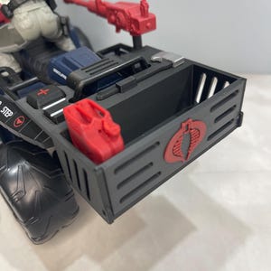 Peut inclure: Un véhicule jouet noir avec un bidon d'essence rouge et un logo Cobra rouge à l'arrière. Le véhicule a un siège noir et bleu et une mitrailleuse rouge sur le dessus.