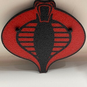 Peut inclure: Un embl&egrave;me de Cobra rouge et noir avec une surface textur&eacute;e. Le Cobra est un symbole de l'organisation fictive Cobra de la franchise G.I. Joe.