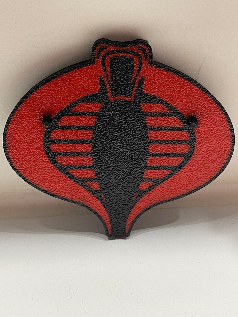 Peut inclure: Un embl&egrave;me de Cobra rouge et noir avec une surface textur&eacute;e. Le Cobra est un symbole de l'organisation fictive Cobra de la franchise G.I. Joe.