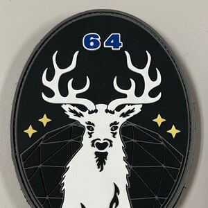 May include: A black and white patch with a white deer with antlers, a black beard, and a geometric pattern in the background. The number 64 is in blue at the top of the patch.
